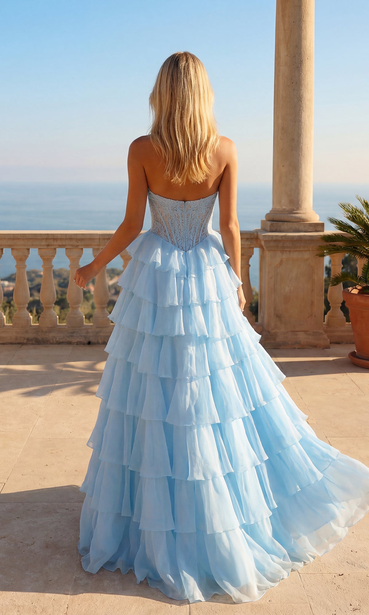 A woman with long blonde hair wears the Long Ruffle Prom Gown: PromGirl Exclusive 250205, a strapless light blue dress, as she stands on a terrace overlooking the sea.
